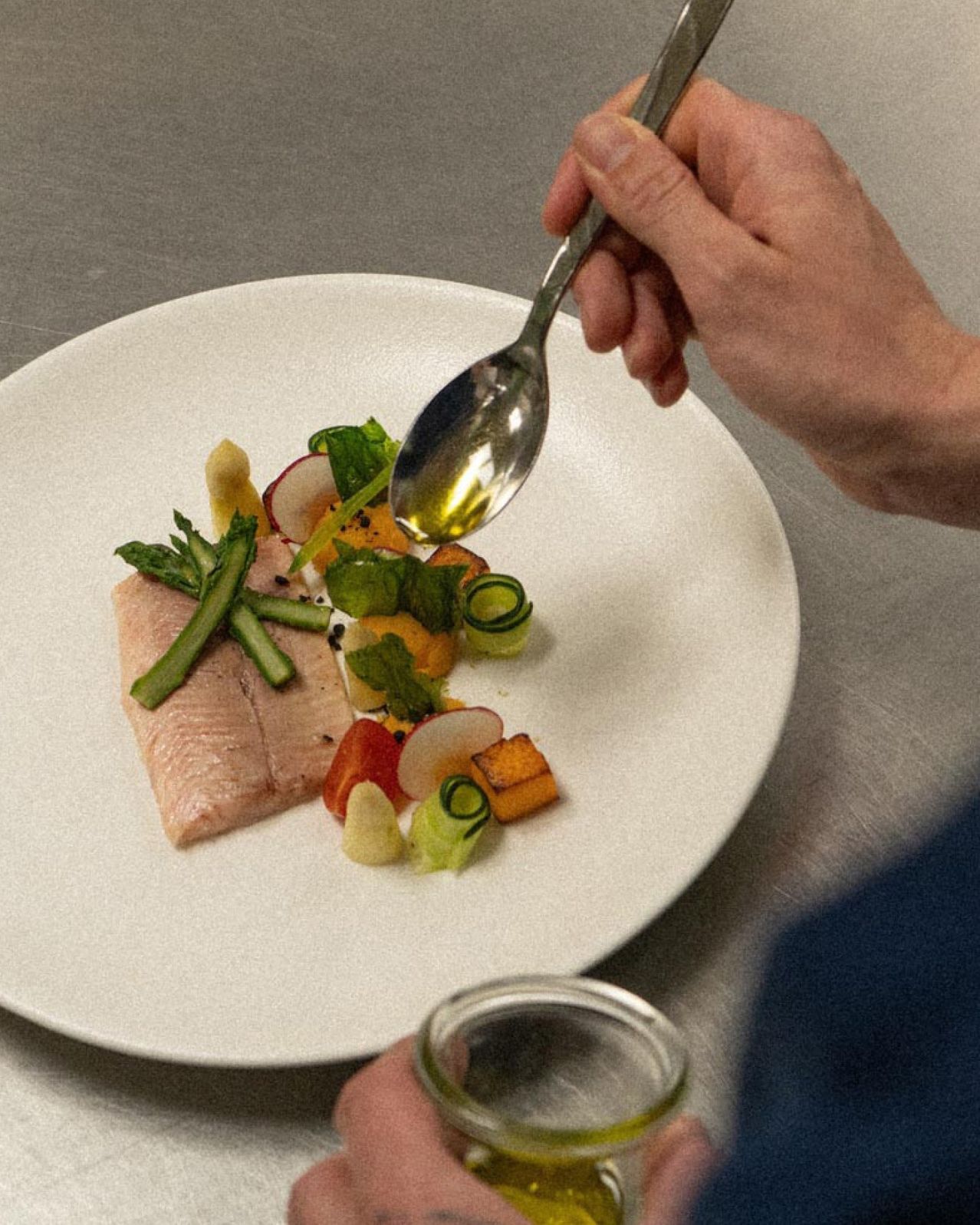 A chef drizzles olive oil over a white plate with fish, vegetables, and herbs.