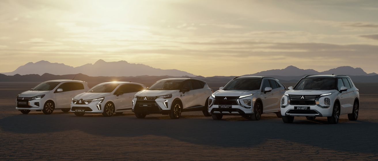 Fünf weiße Mitsubishi SUVs, die in einer Wüstenlandschaft mit Bergen und einer untergehenden Sonne im Hintergrund geparkt sind.