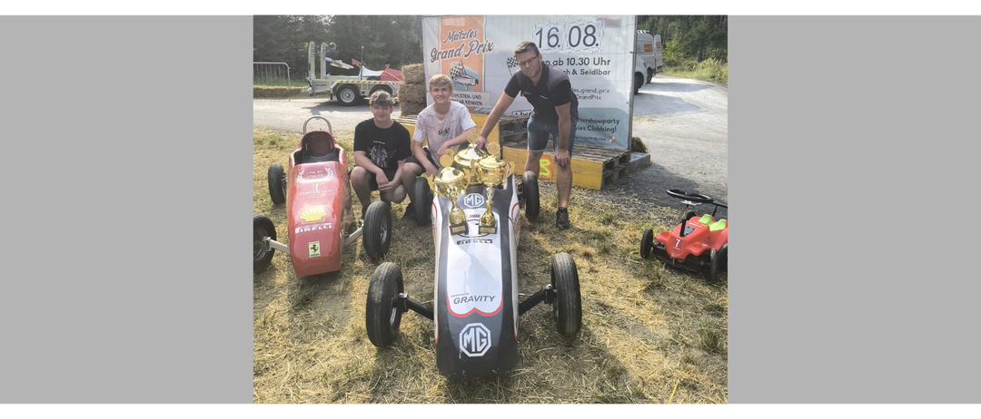 Drei Personen posieren mit Spielzeugautos, einer hält eine Trophäe. Sie befinden sich in einem Außenbereich mit Gras und einem großen Banner.