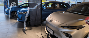 Ein Indoor-Autohaus zeigt eine Auswahl an Autos. Ein weißes Auto mit dem Kennzeichen MGS5 EV steht vorne, dahinter zwei blaue Autos. In der Mitte steht ein Banner.