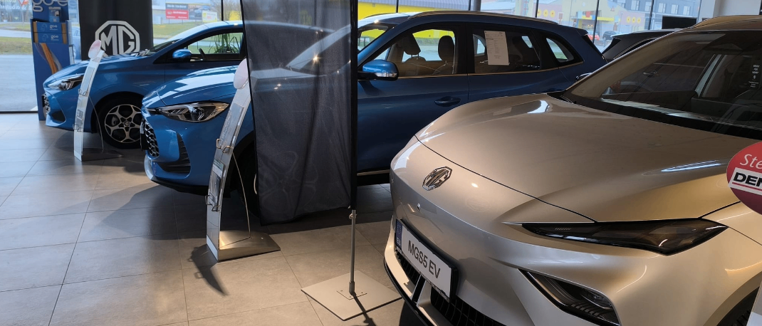 Ein Indoor-Autohaus zeigt eine Auswahl an Autos. Ein weißes Auto mit dem Kennzeichen MGS5 EV steht vorne, dahinter zwei blaue Autos. In der Mitte steht ein Banner.