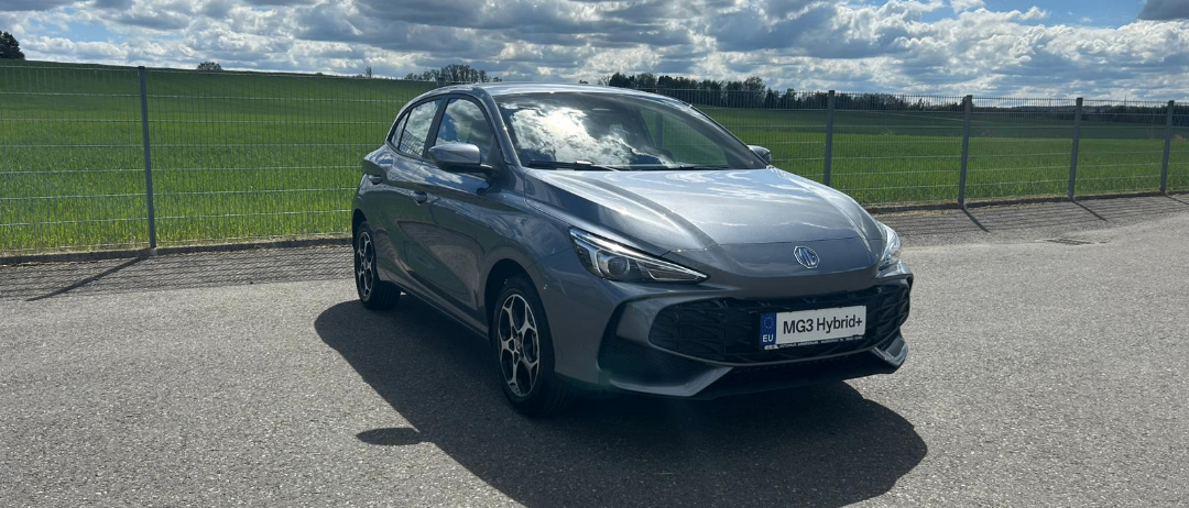 Ein graues MG3 Hybrid+ Auto parkt an einer Straße mit Zaun und grünem Feld im Hintergrund.