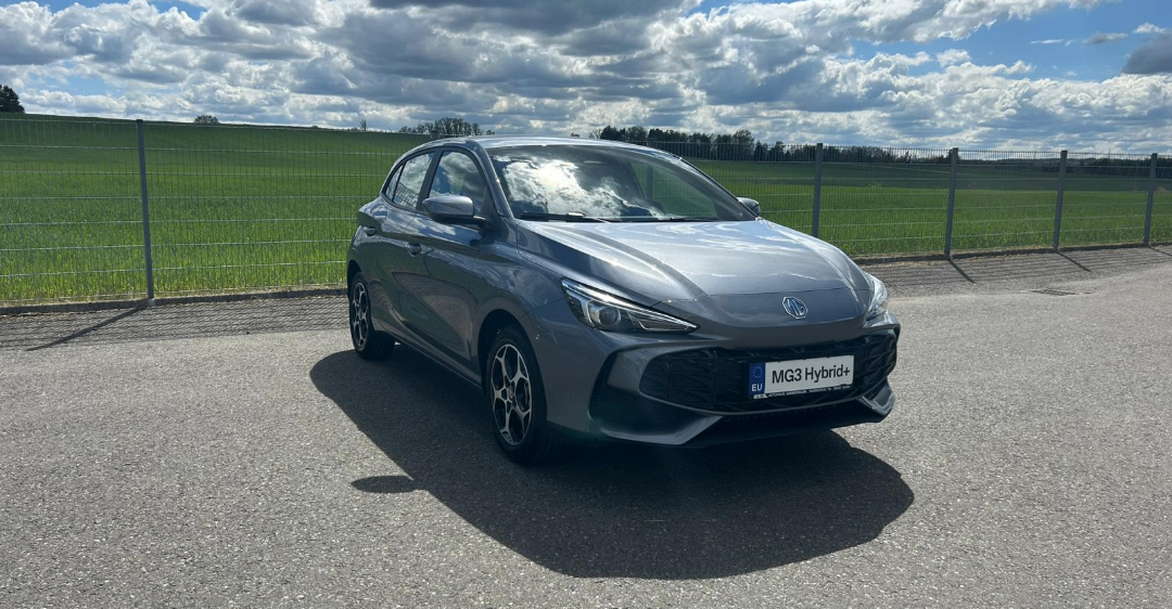 Ein graues MG3 Hybrid+ Auto parkt an einer Straße mit Zaun und grünem Feld im Hintergrund.