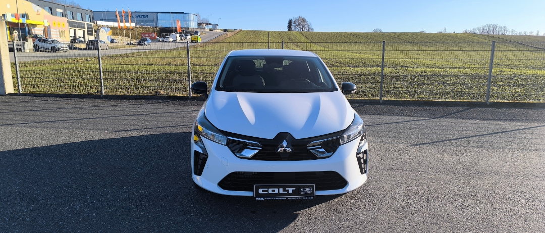 A white Renault Colt is parked in a lot near a fence, with a field and buildings in the background.