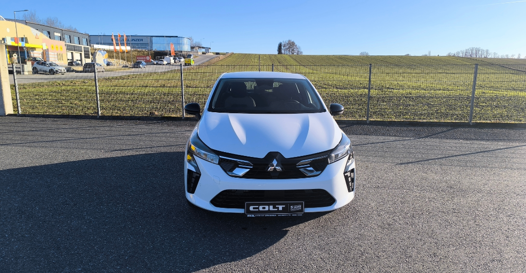 Ein weißer Renault Colt ist in einem Parkplatz in der Nähe eines Zauns geparkt, mit einem Feld und Gebäuden im Hintergrund.