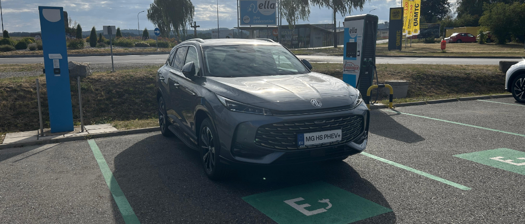 Ein graues Elektroauto ist an einer Ladestation auf einem Parkplatz mit einem Grasbereich und Bäumen im Hintergrund geparkt.