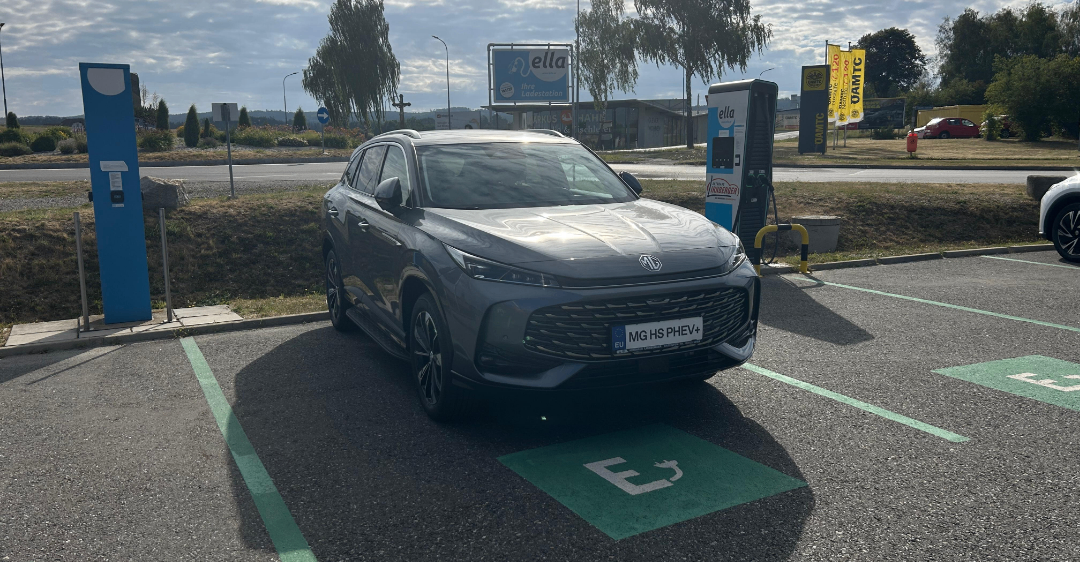 Ein graues Elektroauto ist an einer Ladestation auf einem Parkplatz mit einem Grasbereich und Bäumen im Hintergrund geparkt.
