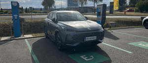 Ein graues Elektroauto ist an einer Ladestation auf einem Parkplatz mit einem Grasbereich und Bäumen im Hintergrund geparkt.