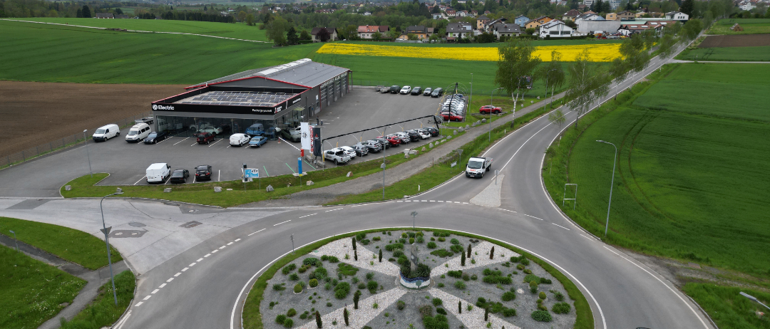 Eine Luftaufnahme eines Autohauses mit mehreren geparkten Autos auf dem Parkplatz und einem Kreisverkehr im Vordergrund.