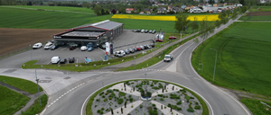 Eine Luftaufnahme eines Autohauses mit mehreren geparkten Autos auf dem Parkplatz und einem Kreisverkehr im Vordergrund.