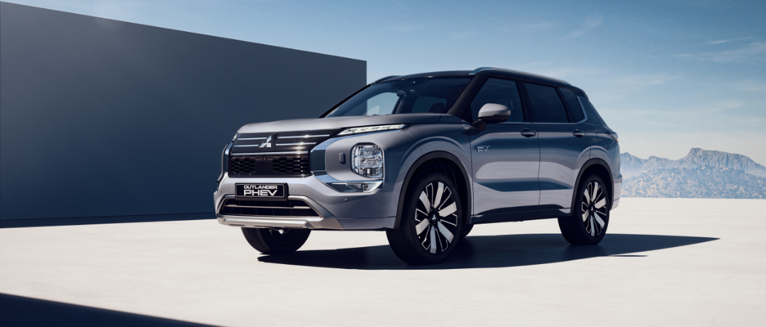 Ein grauer Mitsubishi Outlander PHEV SUV steht vor einer großen Betonmauer mit blauem Himmel und Bergen im Hintergrund.