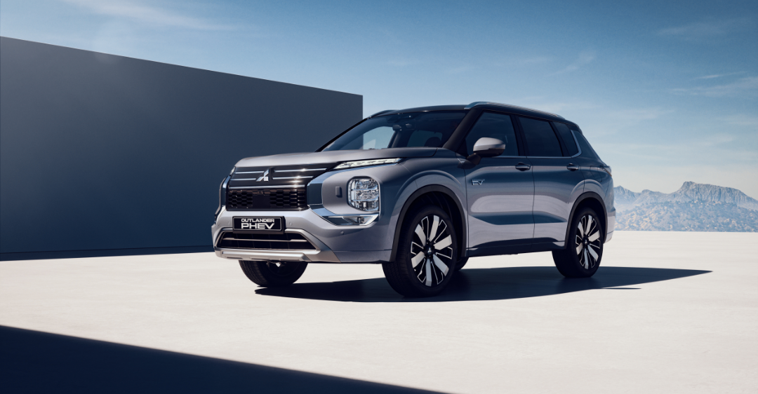 Ein grauer Mitsubishi Outlander PHEV SUV steht vor einer großen Betonmauer mit blauem Himmel und Bergen im Hintergrund.