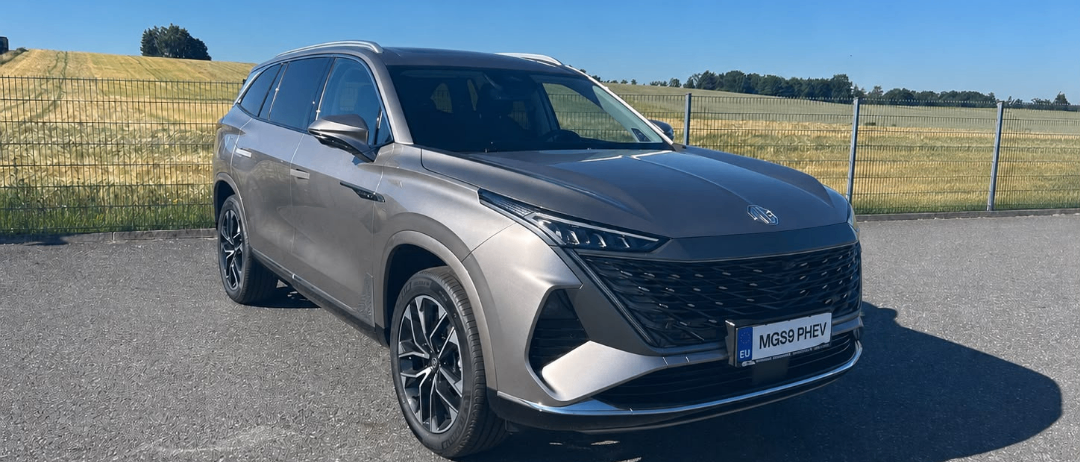 Ein grauer Toyota Highlander SUV mit dem Kennzeichen MGS9 PHEV vor einem Zaun geparkt.