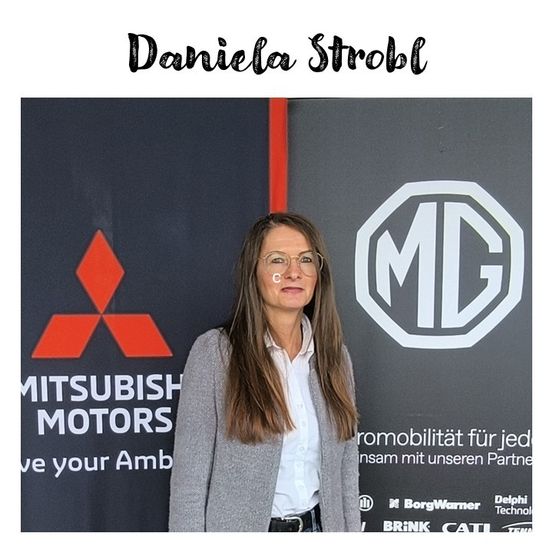 Daniela Strobl steht vor einem Mitsubishi Motors Banner, trägt eine Brille und eine graue Jacke und posiert für ein Foto.