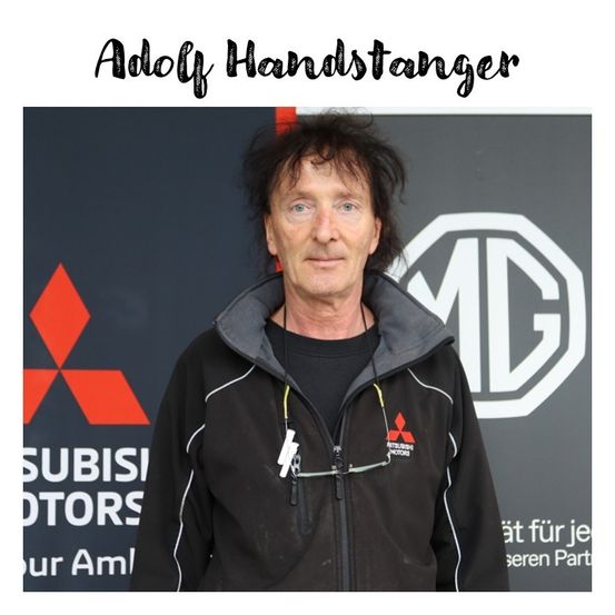 Ein Mann mit langen schwarzen Haaren steht vor einer Wand mit dem Mitsubishi-Logo. Er trägt eine schwarze Jacke mit weißen Streifen und ein schwarzes Unterhemd darunter.