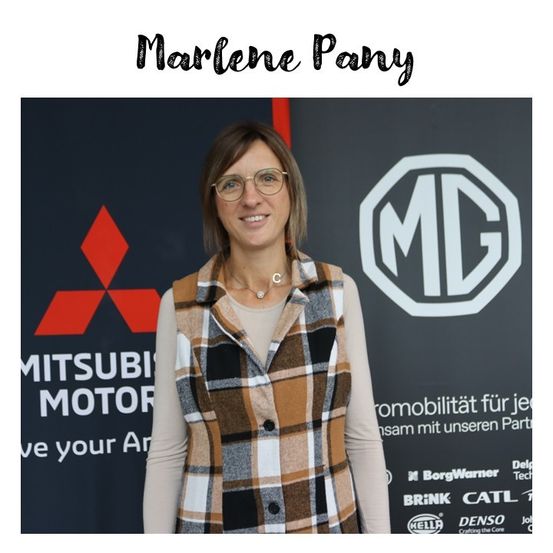 Eine Frau steht vor einem Mitsubishi Motors-Banner und lächelt in die Kamera. Sie trägt eine Brille und eine karierte Weste. Hinter ihr ist das MG-Logo zu sehen.