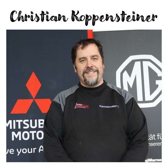 Ein Mann steht vor einem Hintergrund mit Mitsubishi- und MG-Logos. Er trägt ein schwarz-graues Jackett mit einem Logo auf der linken Brust.