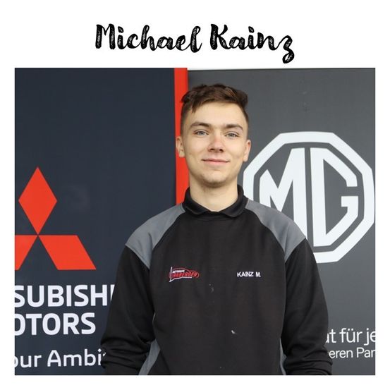 Ein Mann steht vor einer Wand mit Mitsubishi- und MG-Logos und trägt ein schwarz-graues Jackett mit einem roten Logo.