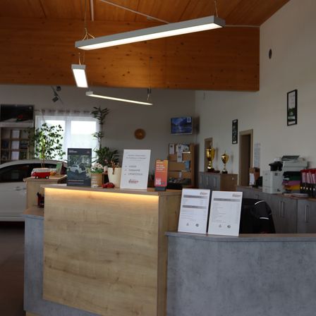 Eine Innenrezeption mit einem hölzernen Schreibtisch, Monitoren, Trophäen, einem Drucker und einem weißen Auto, das draußen geparkt ist. Holzbalken und Lichter sind an der Decke.