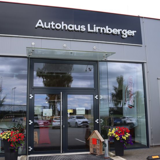 Der Eingang zum Autohaus Limburger ist mit einem großen Glasvordach, Glastüren und einem modernen Schild ausgestattet. Blumenarrangements flankieren den Eingang, und ein kleines Holzhaus steht davor.