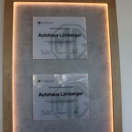 Zwei gerahmte Zertifikate sind an einer Wand angebracht. Das obere Zertifikat lautet 'TOP IN ZUBEHOR 2022' mit dem Namen 'Autohaus Linrberger' und hat Unterschriften darunter. Das untere Zertifikat lautet 'TOP IN AFTER SALES 2023' mit demselben Namen und hat auch Unterschriften. Beide Zertifikate sind von einem Licht beleuchtet.