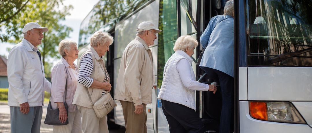 Eine Gruppe älterer Personen, darunter eine Frau in einer weißen Weste, steigen an einem sonnigen Tag in einen Bus ein.
