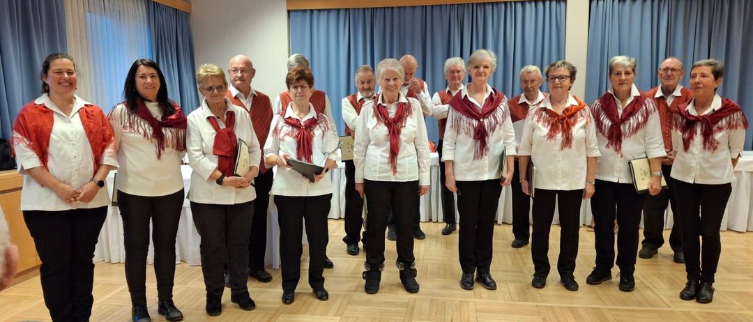 Eine Gruppe von Frauen in weißen Hemden und roten Schals steht in einem Raum mit Holzböden und blauen Vorhängen. Sie halten Bücher und scheinen für ein Foto zu posieren.