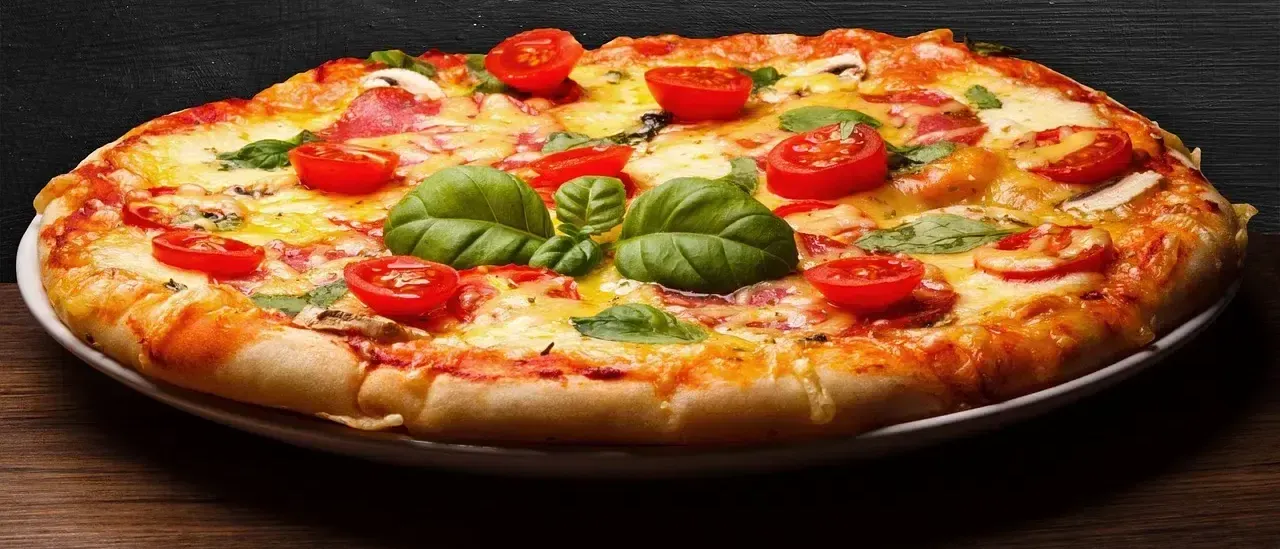 Eine große Pizza mit geschmolzenem Käse, roten Tomaten und Basilikumblättern liegt auf einem schwarzen Teller auf einem Holztisch.