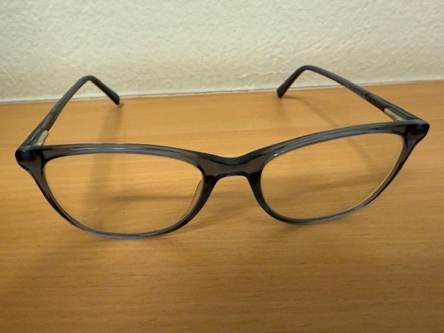 Ein Paar schwarze Brille mit klaren Gläsern liegt auf einem Holztisch. Die Brille hat ein schlankes Design und scheint aus robustem Material zu sein.