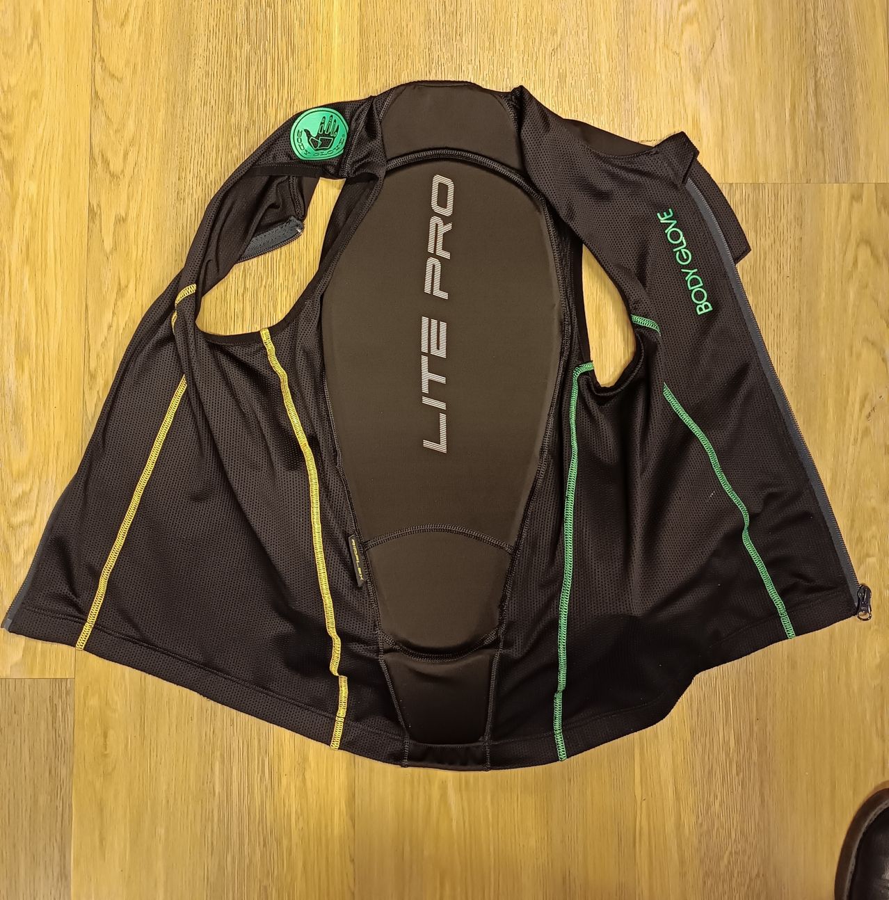 Eine schwarze Body Glove Lite Pro Weste mit grünen und gelben Akzenten liegt flach auf einer Holzoberfläche.