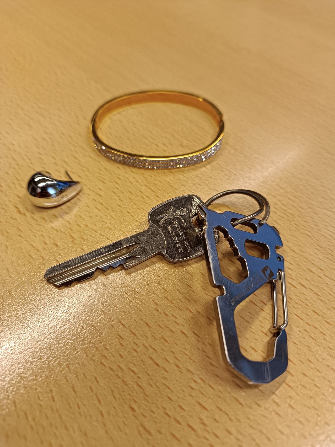 Ein Schlüssel mit einem silbernen Schlüsselanhänger, einem silbernen Ohrring und einer goldenen Armband mit Diamanten auf einer hölzernen Oberfläche.