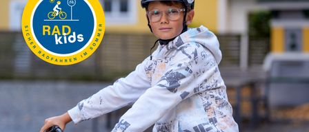 Ein junger Junge fährt mit einem Helm und einer Brille auf einem Fahrrad, mit einem Gebäude und einem Logo im Hintergrund.