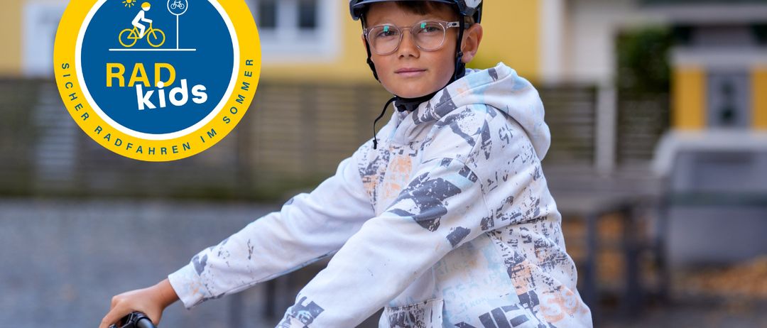 Ein junger Junge fährt mit einem Helm und einer Brille auf einem Fahrrad, mit einem Gebäude und einem Logo im Hintergrund.