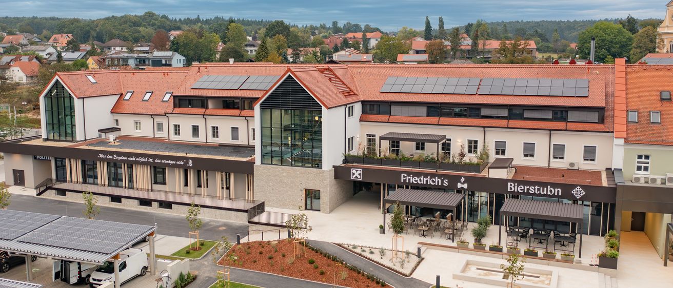 An aerial view of a modern building with a red roof and solar panels, surrounded by trees and a clear blue sky. The building has glass windows, a large entrance, and a sign that reads 'Friedrich's Bierstube'.