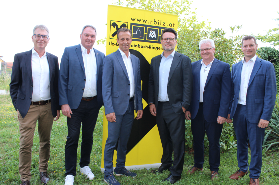 Fünf Männer in Anzügen posieren für ein Foto vor einem gelben Banner mit der Website www.rbilz.at.