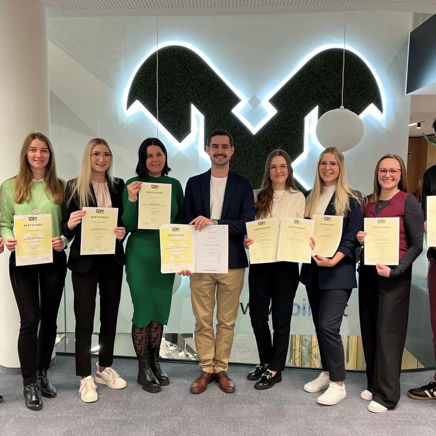 Eine Gruppe von acht Menschen, alle lächelnd, steht vor einer Glaswand mit einem Logo. Sie halten Zertifikate in den Händen, und einige tragen Brillen und Halsketten. Die Umgebung ist in Innenräumen.