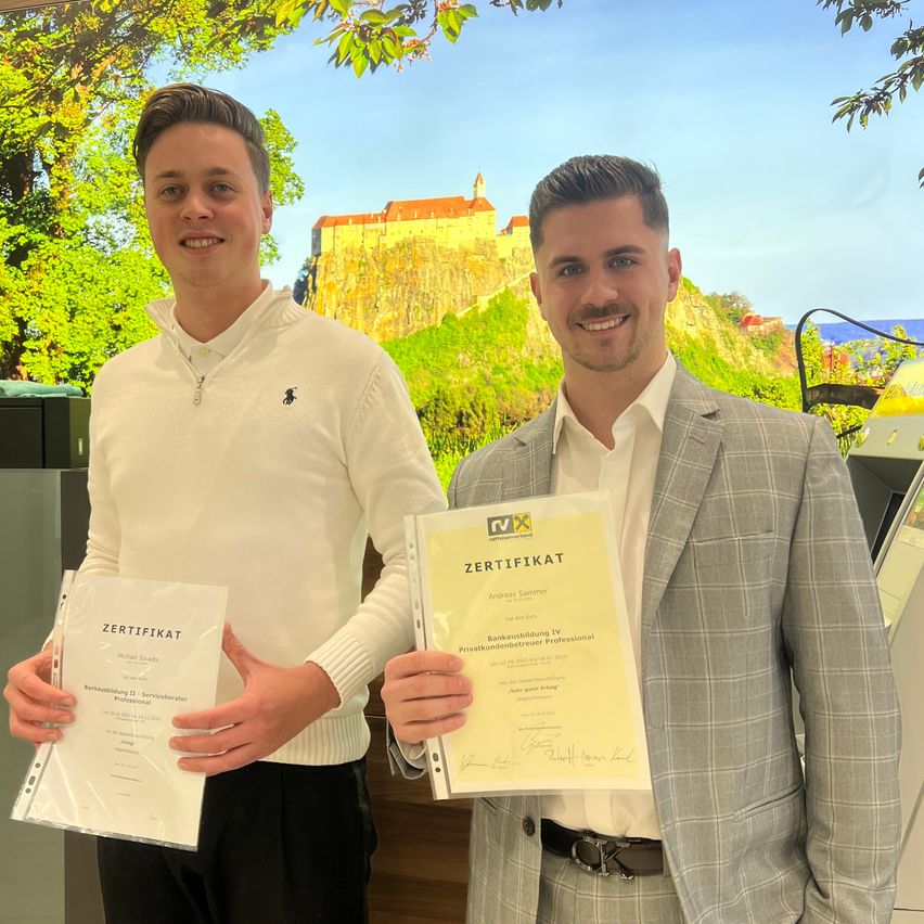 Zwei Männer lächeln und posieren für ein Foto, während sie Zertifikate halten. Sie stehen vor einer Kulisse aus Burg und Grün. Einer hält ein weißes Zertifikat, der andere ein gelbes.