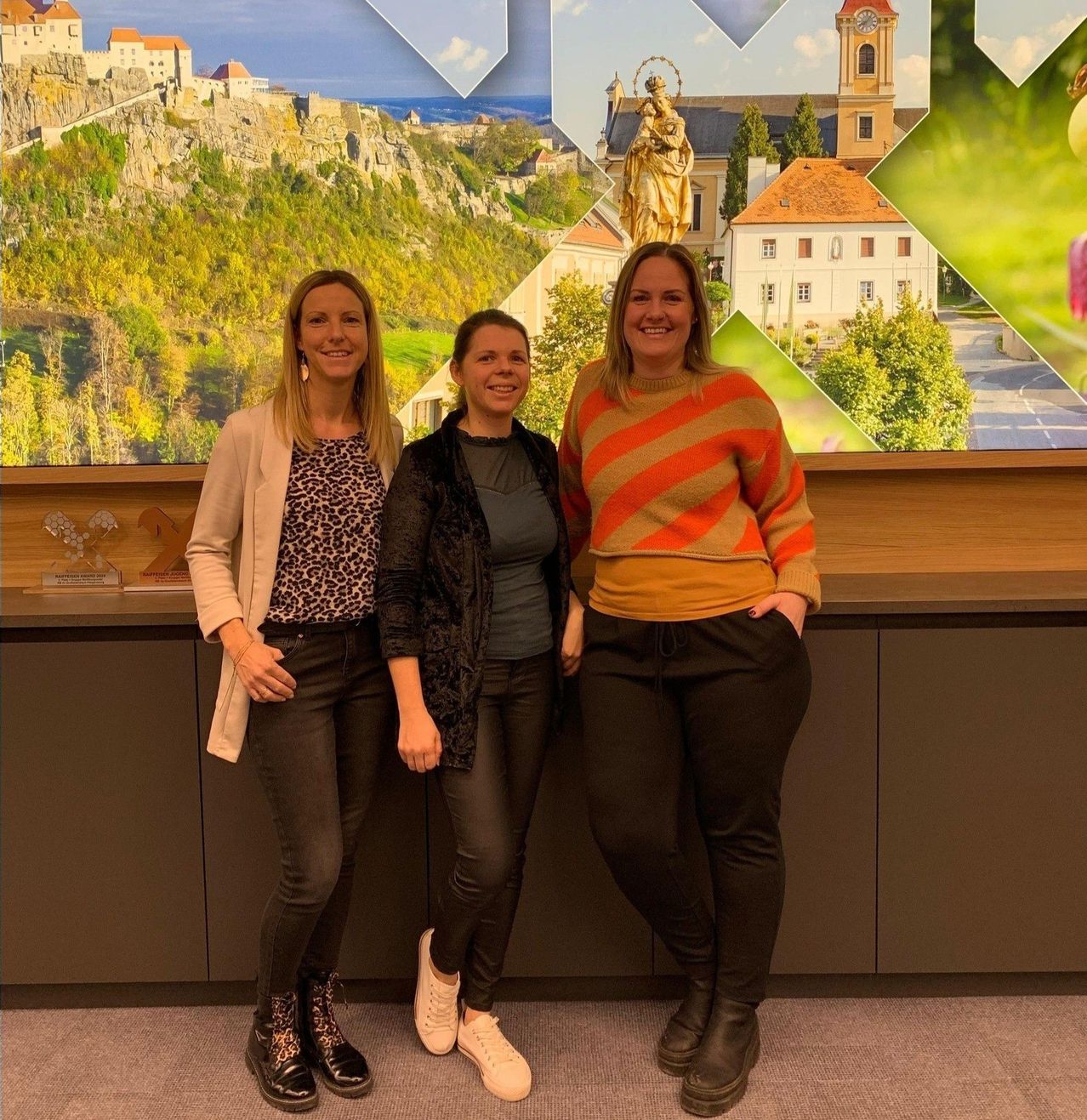 Drei Frauen stehen gemeinsam vor einem großen Wandbild, das eine malerische Landschaft mit Gebäuden und einer Statue zeigt.