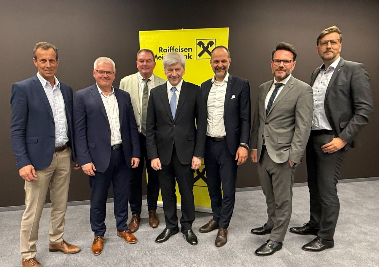 Six men in formal attire standing in front of a yellow banner with 'Raiffeisen' written on it.
