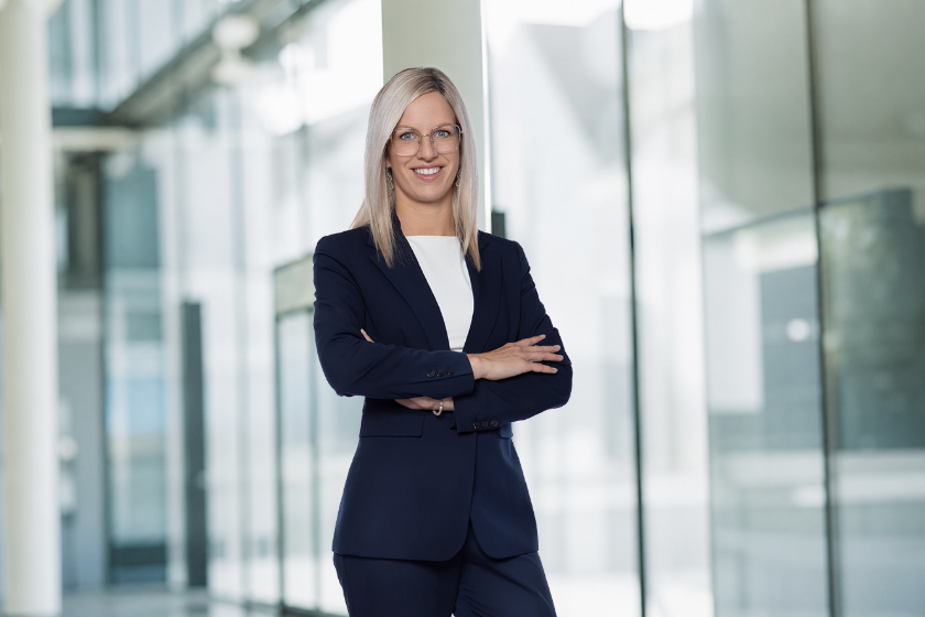 A woman in a navy blue suit stands confidently with her arms crossed in a modern office building.