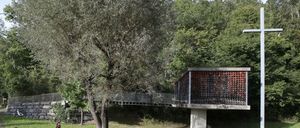 Eine Gruppe von Menschen sitzt im Kreis auf Bänken in einem Park. Eine Frau steht und spricht mit einer anderen Frau. Eine Brücke ist im Hintergrund.