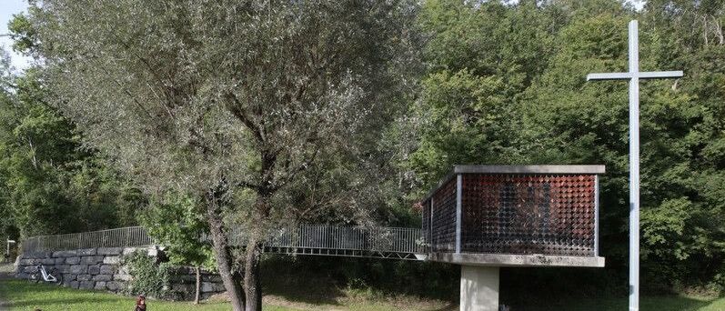 Eine Gruppe von Menschen sitzt im Kreis auf Bänken in einem Park. Eine Frau steht und spricht mit einer anderen Frau. Eine Brücke ist im Hintergrund.