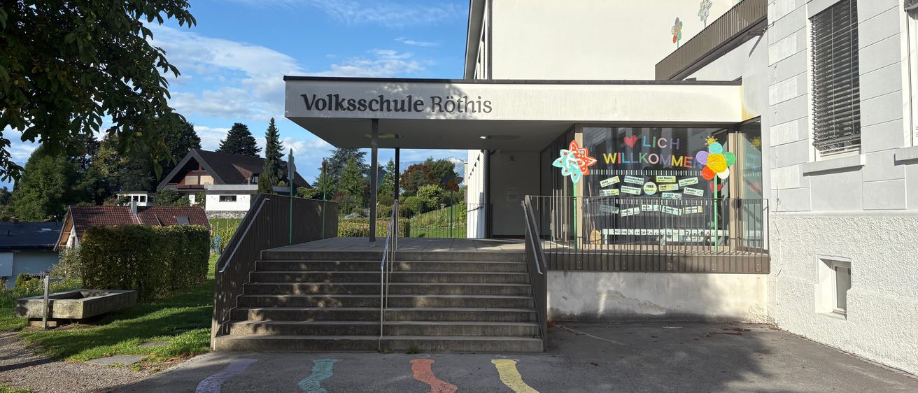 The Volksschule Rothis school building has a large entrance with stairs leading up. A sign reads 'Volksschule Rothis'. A decorative window displays colorful decorations. A house and trees are in the background.