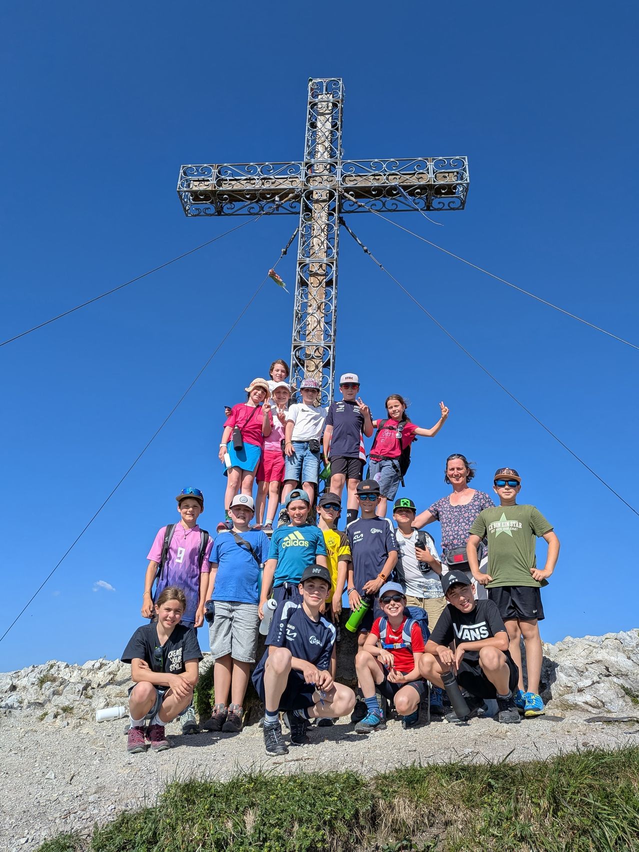 Eine Gruppe von Kindern und Erwachsenen posiert für ein Foto vor einem großen Kreuz auf einem Berg unter einem klaren blauen Himmel.