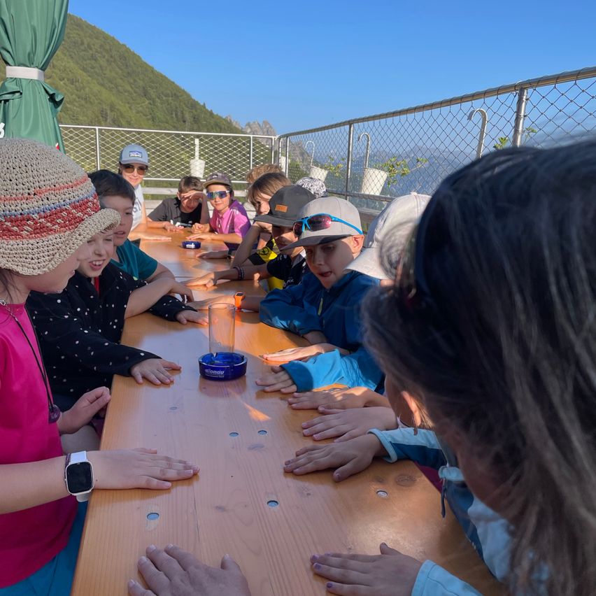Eine Gruppe von Kindern mit Hüten sitzt an einem Tisch und ist in eine Aktivität vertieft. Ein Kind konzentriert sich auf einen blauen Gegenstand auf dem Tisch. Sie befinden sich in einer Outdoor-Umgebung mit Bergen und einem klaren blauen Himmel im Hintergrund.