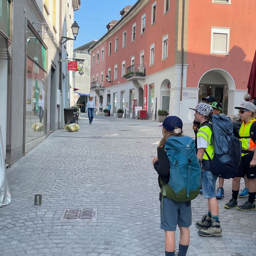Eine Gruppe von Kindern mit Rucksäcken und Sicherheitswesten steht auf einer Kopfsteinpflasterstraße und blickt zu einem Gebäude hinauf. Eine Frau geht im Hintergrund.