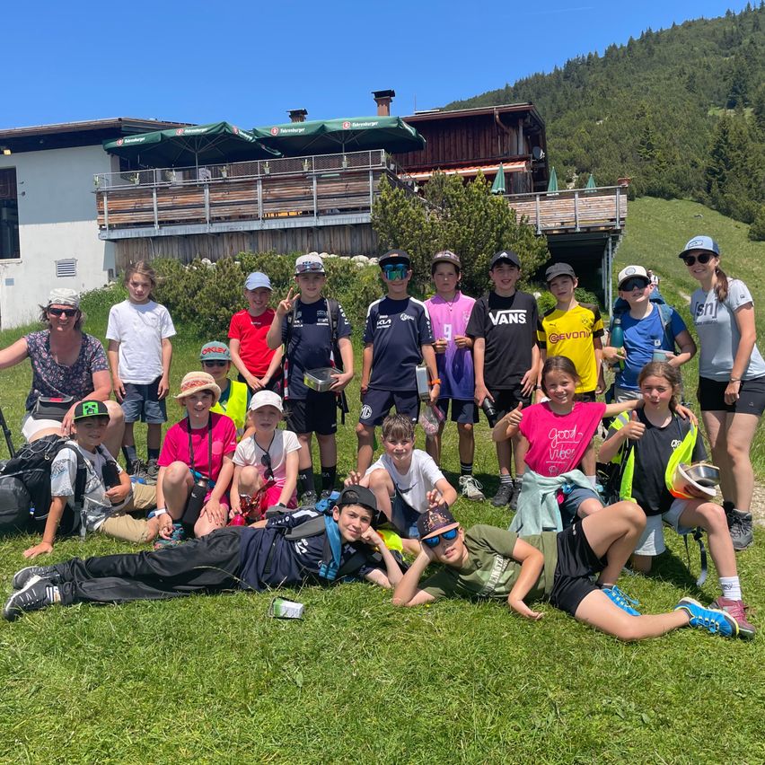 Eine Gruppe junger Leute in Mützen und Turnschuhen, einige mit Helmen, posiert für ein Foto vor einem Gebäude mit Balkon und Sonnenschirmen.