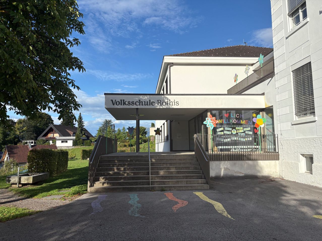 Der Haupteingang der Volksschule Rothis, ein Schulgebäude mit Stufen, die zum Eingang führen. Das Gebäude hat ein Schild über dem Eingang und bunte Dekorationen am Glasfenster.