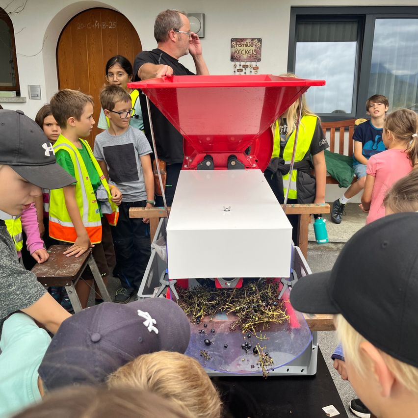 Eine Gruppe von Kindern und einem Erwachsenen beobachten eine rote und weiße Maschine, die möglicherweise zur Verarbeitung von etwas verwendet wird. Die Kinder tragen Sicherheitswesten und Mützen. Hinter ihnen befindet sich eine Holztür, ein Fenster und eine Bank.