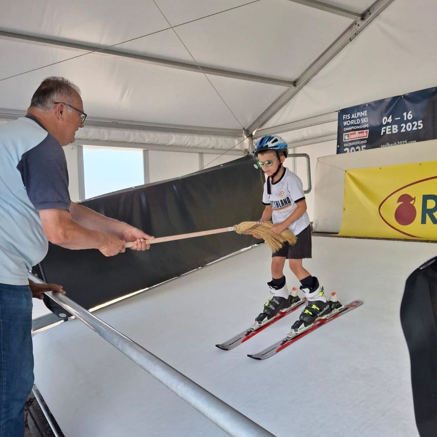 Ein erwachsener Mann kehrt den Schnee, während ein Kind auf einer Indoor-Ski-Piste Ski fährt, wahrscheinlich bei einer Veranstaltung.
